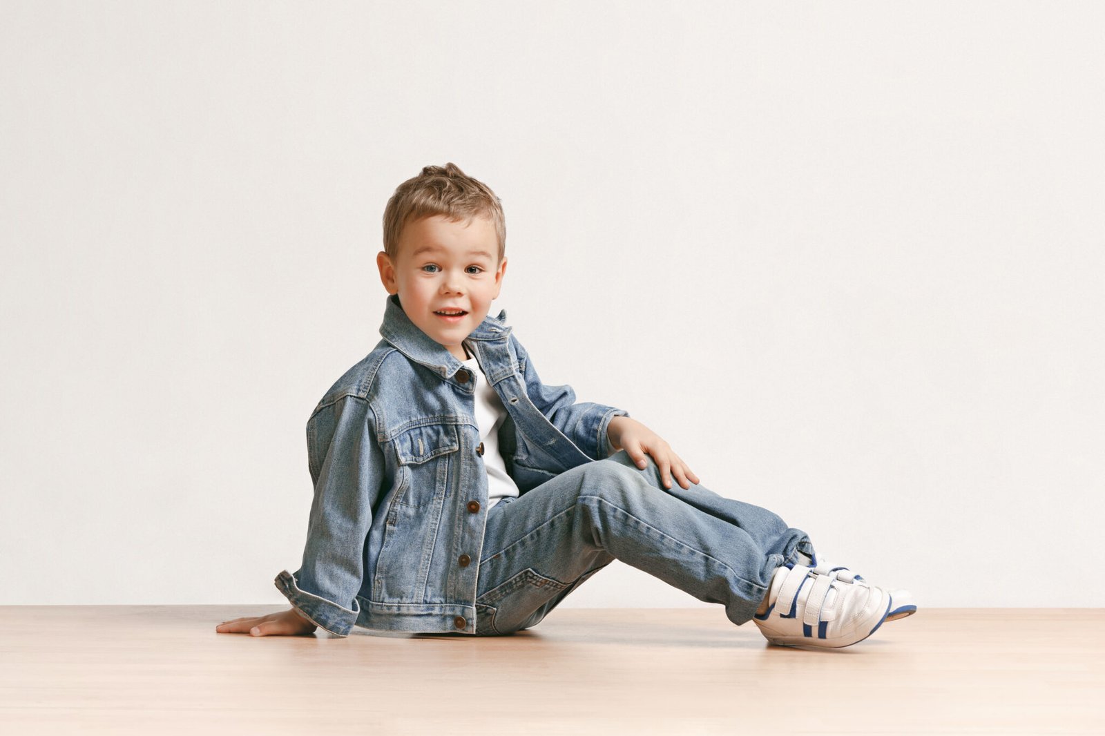 The portrait of cute little kid boy in stylish jeans clothes looking at camera against white studio wall. Kids fashion concept