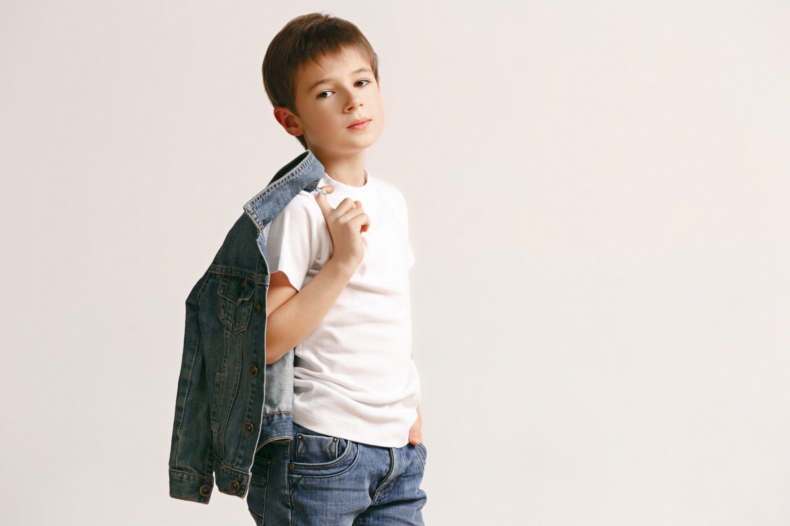 The portrait of cute little kid boy in stylish jeans clothes looking at camera against white studio wall. Kids fashion concept